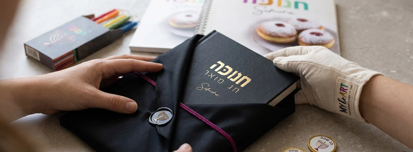 המדריך המלא למנהלי רווחה: איך לבחור מתנות לעובדים שבאמת יעריכו ב-2026 6 קלוזאפ פוטו-ריאליסטי על ידיים של עובדת פותחת בעדינות אריזת משי שחורה. מתוך האריזה מבצבצת כריכה של יומן יוקרתי עם הטבעת לוגו מוזהבת ועדינה. עומק שדה רדוד המדגיש את איכות החומרים.