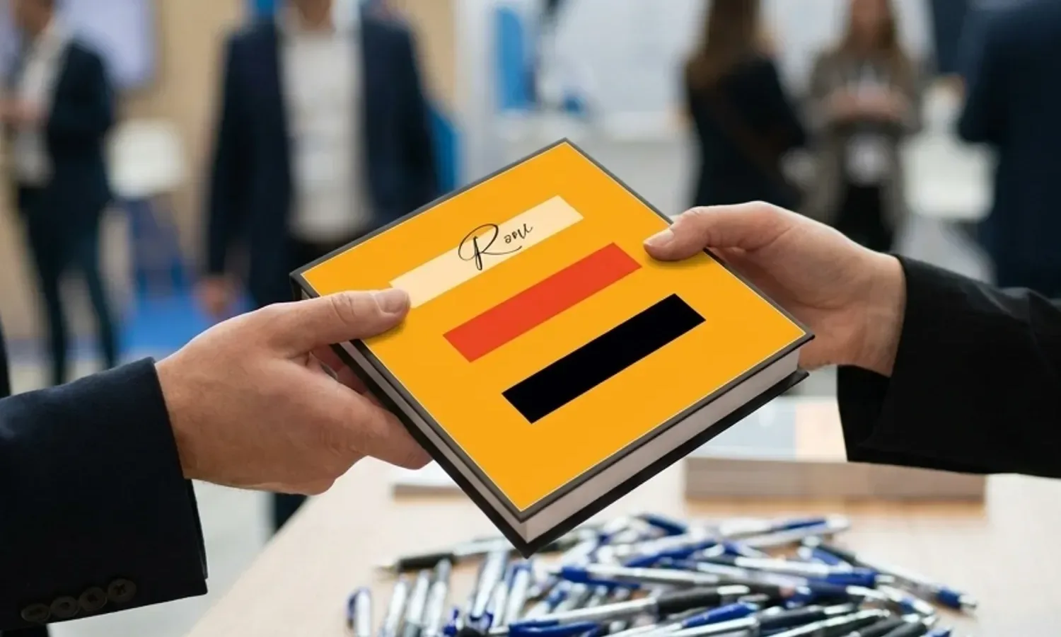 יד של משתתף בכנס מקבלת בביתן תצוגה מחברת מעוצבת ואיכותית, בניגוד לערימת עטי פלסטיק זולים ברקע.